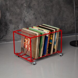 May include: A red metal record storage cart on wheels, filled with vinyl record albums. The cart has a rectangular frame and a wire support. The albums have various colorful covers, and the cart is on a gray surface.