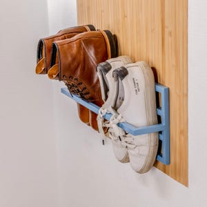 May include: A blue metal shoe rack mounted on a light wood panel, holding two pairs of shoes. The shoes include brown leather boots and white sneakers. The rack is attached to a white wall.