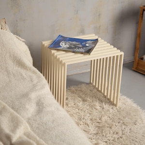 May include: A light-colored wooden side table with a slatted design. A magazine with a blue cover rests on top. The table is placed on a shaggy, cream-colored rug, next to a soft blanket.