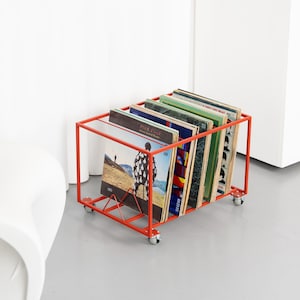 Könnte beinhalten: Ein roter Metall-Schallplattenwagen auf Rädern, gefüllt mit Vinyl-Schallplatten. Der Wagen hat einen rechteckigen Rahmen und ein Drahtgestelldesign. Die Albumcover sind sichtbar und zeigen verschiedene Albumkunst und -titel.