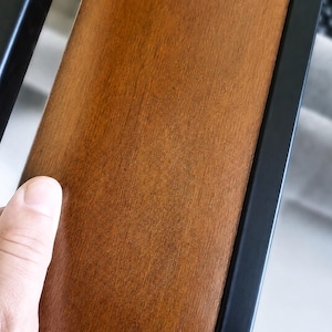 May include: A smooth, medium-brown wooden panel set between two vertical matte black metal bars. The wood grain is visible across the surface, showing a warm, polished finish. The structure appears to be part of a larger furniture piece or railing.