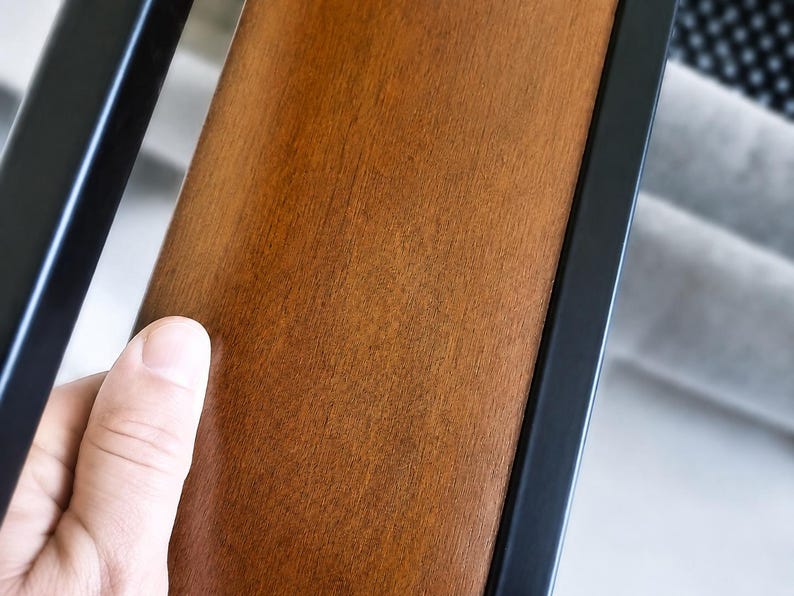 May include: A smooth, medium-brown wooden panel set between two vertical matte black metal bars. The wood grain is visible across the surface, showing a warm, polished finish. The structure appears to be part of a larger furniture piece or railing.