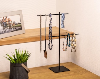 Könnte beinhalten: Ein schwarzer Schmuckständer aus Metall mit verschiedenen Halsketten und Armbändern. Zu den Halsketten gehören eine silberne Kette, eine blaue Perlenkette und eine Anhängerkette. Die Armbänder sind mit bunten Perlen verziert. Der Ständer steht auf einer Holzoberfläche.