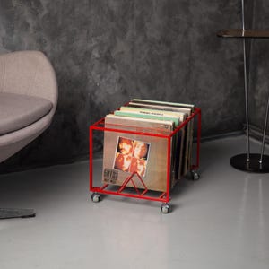 Może przedstawiać: Czerwony metalowy wózek do przechowywania płyt winylowych na kółkach, wypełniony albumami. Okładki albumów przedstawiają zdjęcia muzyków i tekst. Wózek znajduje się w pomieszczeniu z szarą ścianą i podłogą.