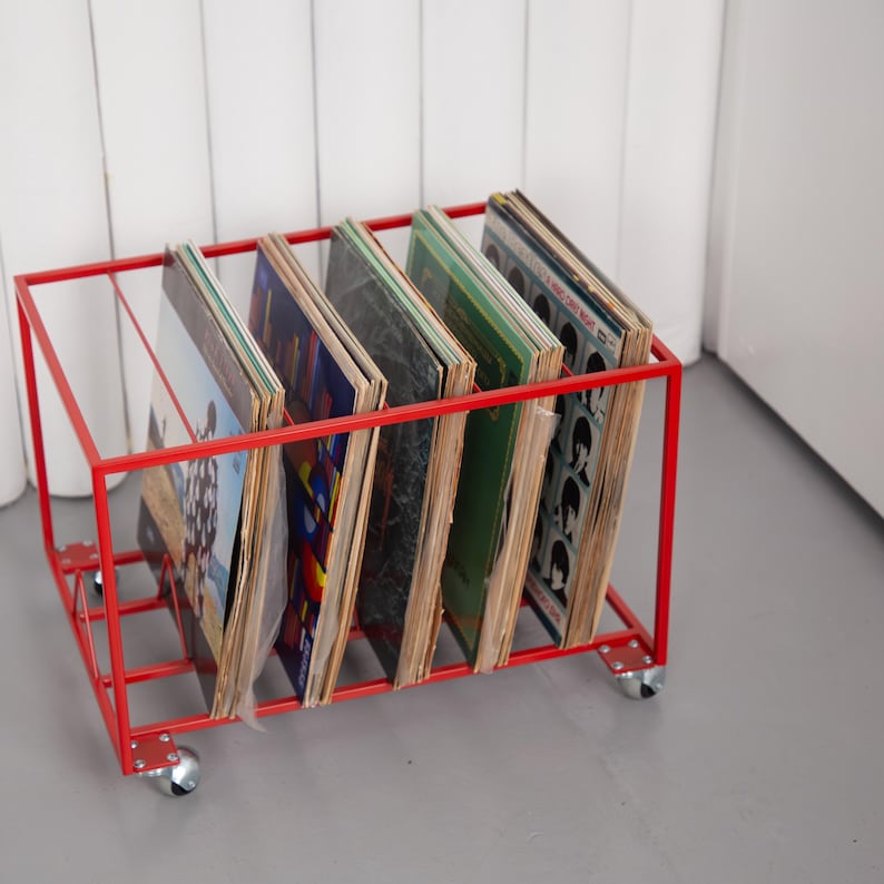 May include: A red metal record storage rack on wheels, holding several vinyl record albums. The rack has a rectangular frame and is designed to display and organize records. The albums have various colorful covers.
