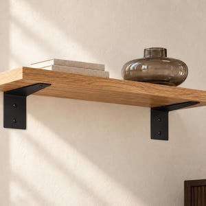 May include: A light brown wooden floating shelf mounted with two black metal L-shaped brackets. On the shelf, there is a stack of two light-colored books and a round, translucent, dark-tinted glass vase with a wide base and a narrow neck.
