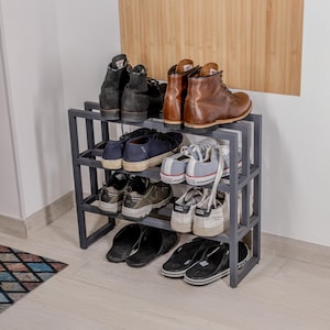Könnte beinhalten: Ein graues Schuhregal aus Metall, gefüllt mit verschiedenen Schuhen. Zu den Schuhen gehören schwarze Stiefel, braune Lederstiefel, Turnschuhe und Sandalen. Das Regal hat drei Ebenen und steht vor einer weißen Wand.