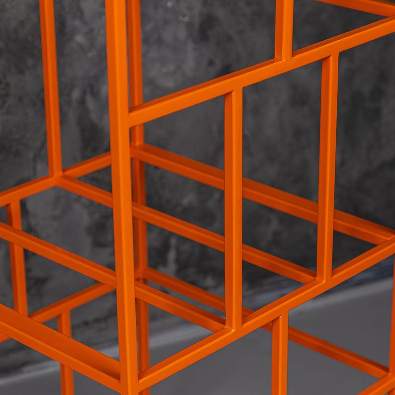 May include: Close-up of an orange metal geometric structure. The structure is made of straight lines and right angles, creating a grid-like pattern. The metal has a matte finish. The background is a dark gray, textured wall.