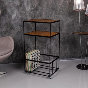 May include: A black metal and wood record storage unit with three shelves. The top and middle shelves are wood, and the bottom shelf is metal. A stack of vinyl records is stored on the bottom shelf. The unit has wheels.