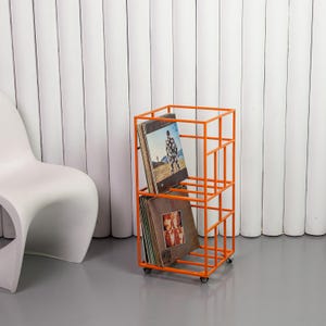 May include: An orange metal record storage rack on wheels, holding two vinyl records. The rack has a geometric, open design. The background features a white chair and a white, vertically-lined wall.