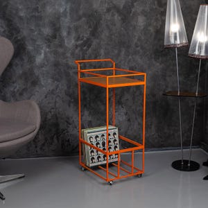 May include: An orange metal record cart with a wooden shelf and a lower compartment holding vinyl records. The cart has a rectangular frame and sits on small wheels. The background features a grey wall and a grey floor.