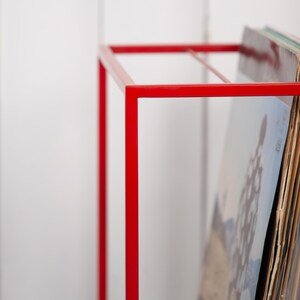 May include: A bright red, minimalist record storage rack. The metal frame is a simple geometric design, holding a stack of vinyl records. The rack's bold color contrasts with the white background, highlighting the records.