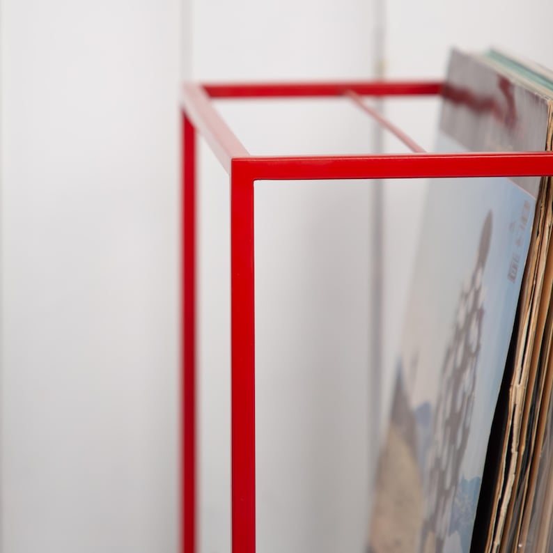 May include: A bright red, minimalist record storage rack. The metal frame is a simple geometric design, holding a stack of vinyl records. The rack's bold color contrasts with the white background, highlighting the records.
