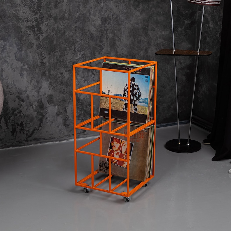 May include: An orange metal record storage unit on wheels. The unit has a rectangular frame with multiple compartments holding vinyl records. The records have album art visible, and the unit is on a gray floor.