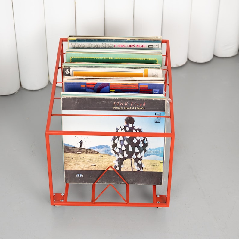 May include: A bright red metal record holder filled with vinyl records. The holder has a geometric design and is filled with album covers, including Pink Floyd's "Delicate Sound of Thunder" and The Beatles' "A Hard Day's Night".
