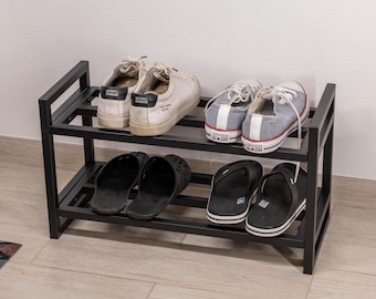 May include: A black metal shoe rack with two shelves. The top shelf holds white sneakers and blue and white striped canvas shoes. The bottom shelf displays black sandals and flip-flops. The rack is on a light wood floor.