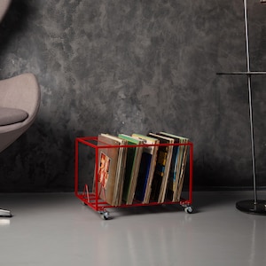 May include: A red metal record storage cart on wheels, filled with vinyl records. The cart has a rectangular frame and is positioned on a gray floor. The records have various album covers.