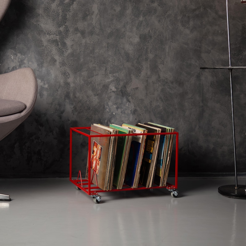 May include: A red metal record storage cart on wheels, filled with vinyl records. The cart has a rectangular frame and is positioned on a gray floor. The records have various album covers.