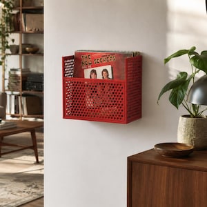 Peut inclure: Un support de rangement mural en métal rouge avec un motif perforé en nid d'abeille. Le support contient un disque vinyle dont la pochette porte le texte The New Seekers. L'unité rectangulaire est fixée à un mur blanc pour l'exposition.