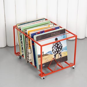 May include: A red metal record storage cart filled with vinyl records. The cart has a rectangular frame with wheels and a triangular support. The album covers are visible, including one by Pink Floyd.