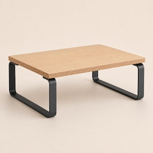 May include: A rectangular monitor stand featuring a light brown wood grain top surface with visible layered plywood edges. The stand is supported by two matte black metal legs that curve into a U-shape on each side, providing a stable base.