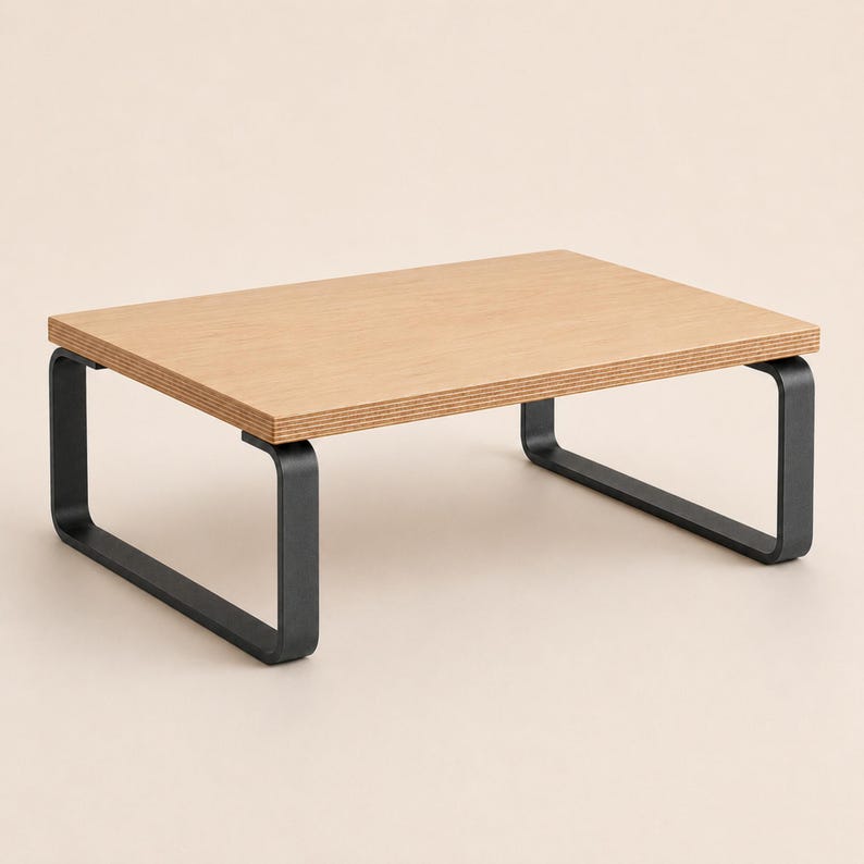 May include: A rectangular monitor stand featuring a light brown wood grain top surface with visible layered plywood edges. The stand is supported by two matte black metal legs that curve into a U-shape on each side, providing a stable base.