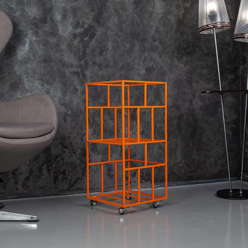May include: An orange metal shelving unit with a geometric design. The unit has multiple shelves and is mounted on small wheels. The background features a gray wall and a gray floor.
