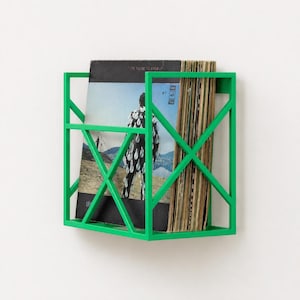 Può includere: Una mensola da parete in metallo verde brillante con un telaio geometrico a forma di X. La mensola contiene una collezione di dischi in vinile, tra cui uno con una copertina visibile che mostra una persona con un abito a motivi.