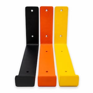 May include: Three L-shaped metal shelf brackets in black, orange, and yellow. Each bracket features a vertical back plate with two circular mounting holes and a horizontal base with a small upturned lip at the front edge for shelf support.