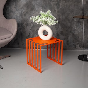 May include: A vibrant orange metal side table with a distinctive geometric design. A white ceramic vase with a circular opening holds white flowers. The table is a modern accent piece, suitable for a living room or bedroom.