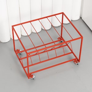 May include: A red metal storage rack with a rectangular frame and angled dividers. The rack sits on four small caster wheels. The design is simple and modern, suitable for organizing items.