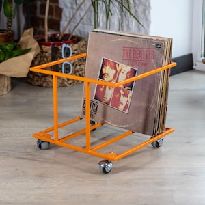 Op de afbeelding: Een oranje metalen platenkar op wielen bevat verschillende vintage vinylplaten. De albums zitten in een bruine hoes met de tekst "THE BEATLES A TASTE OF HONEY" en afbeeldingen van de bandleden. De kar heeft een eenvoudig, rechthoekig ontwerp.