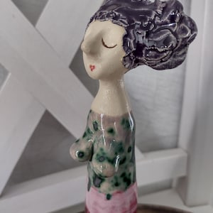 Könnte beinhalten: Eine Keramikfigur einer Person mit geschlossenen Augen und dunkelviolettem Haar. Die Figur hat ein cremefarbenes Gesicht, einen Oberkörper mit grün-violetter Sprenkelglasur und einen unteren Bereich mit rosa und dunkelroten Tropfenmustern.