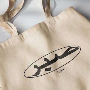 May include: A beige tote bag with a black oval design featuring Arabic calligraphy and the word "Sabr" printed below.