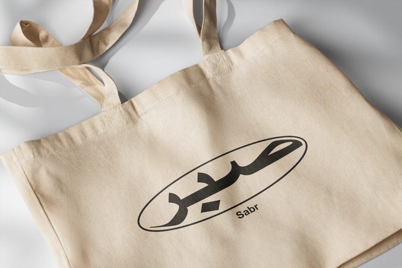 May include: A beige tote bag with a black oval design featuring Arabic calligraphy and the word "Sabr" printed below.