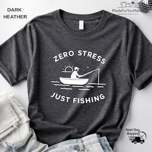 May include: Dark heather gray t-shirt with the text "ZERO STRESS JUST FISHING" in white. The graphic shows a person fishing from a boat. The shirt is made of a soft material.