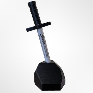 May include: A black 3D printed sword-shaped pen holder with a white Sharpie marker inserted. The marker has the text "Extra Fine Point Permanent Marker" on it.