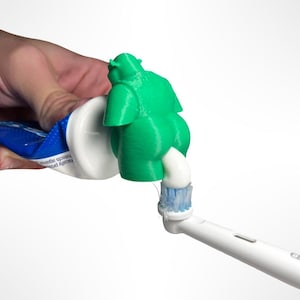 Puede incluir: Un soporte verde con forma de Shrek para el tubo de pasta de dientes que dispensa pasta dental blanca sobre un cepillo de dientes blanco y azul. El soporte está impreso en 3D y añade un toque divertido a la decoración del baño.