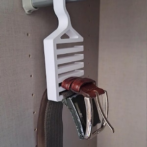 May include: A white plastic belt hanger with multiple slots, hanging from a closet rod. Several belts of different colors and styles are hanging from the slots, showcasing a storage solution for accessories.