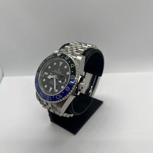Peut inclure: Une montre-bracelet en acier inoxydable avec un cadran noir et une lunette bleue et noire. La montre a un bracelet argenté et est présentée sur un présentoir noir. Le cadran de la montre comporte des index et des aiguilles blancs.