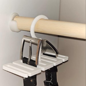 May include: White belt hanger with two black leather belts hanging from it. The hanger is attached to a wooden rod inside a closet. The belts have silver buckles.