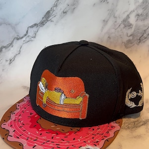 Könnte beinhalten: Schwarze Baseballkappe mit einem gestickten Bild einer Figur auf einem Sofa. Der Schirm der Kappe ist wie ein rosa Donut mit Streuseln und einem Biss gestaltet. Die Kappe hat ein Logo auf der Seite.