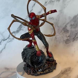 May include: A detailed figurine of Spider-Man in a dynamic pose, clad in a red and gold suit with black accents. The figure is mounted on a detailed, textured base, and features multiple golden mechanical arms extending from the back.