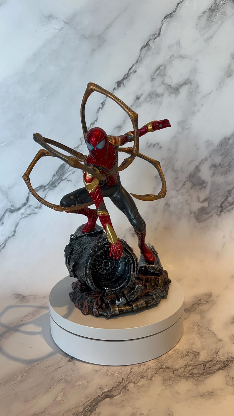 May include: A detailed figurine of Spider-Man in a dynamic pose, clad in a red and gold suit with black accents. The figure is mounted on a detailed, textured base, and features multiple golden mechanical arms extending from the back.
