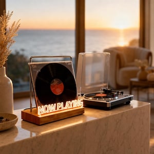 May include: A wooden display stand featuring a clear acrylic holder for a vinyl record, illuminated by a bright white neon sign that reads NOW PLAYING in capital letters, positioned next to a silver and black turntable on a marble surface.