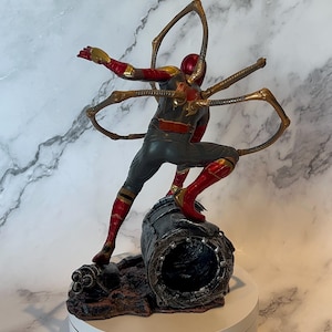 May include: A detailed figurine of a superhero in a dynamic pose, clad in a red, gold, and grey suit. The figure is mounted on a textured base, with metallic gold appendages extending from the back. The figurine is approximately 20 cm tall.