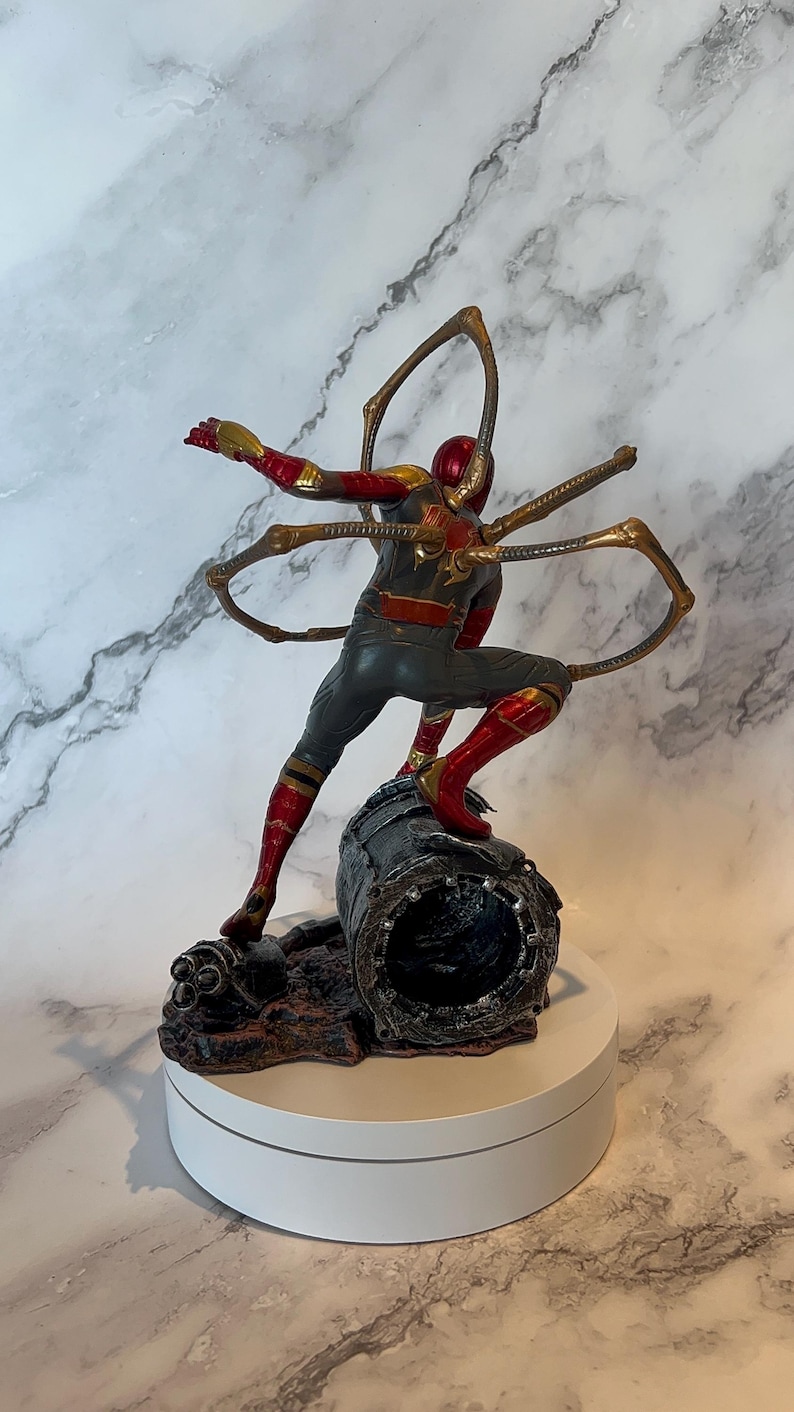 May include: A detailed figurine of a superhero in a dynamic pose, clad in a red, gold, and grey suit. The figure is mounted on a textured base, with metallic gold appendages extending from the back. The figurine is approximately 20 cm tall.