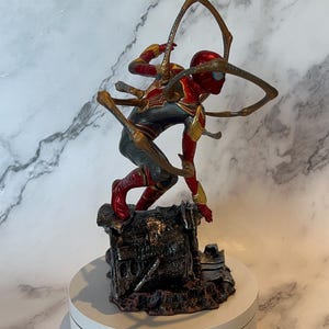 May include: A detailed figurine of Spider-Man in a dynamic pose, clad in a red, gold, and grey suit. The figure is mounted on a detailed, textured base and features multiple mechanical arms extending from the back. The figurine is displayed on a white rotating platform.