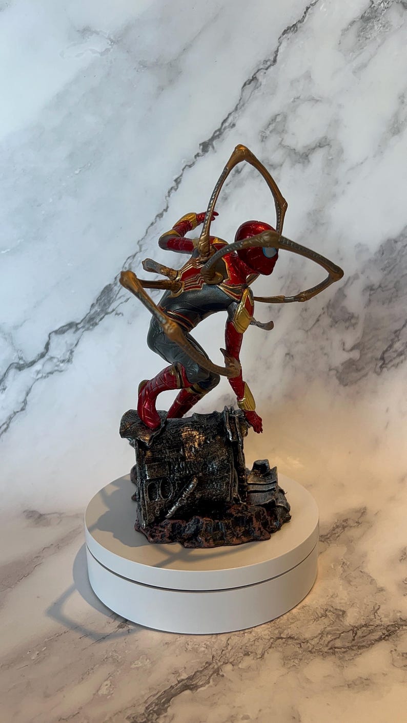 May include: A detailed figurine of Spider-Man in a dynamic pose, clad in a red, gold, and grey suit. The figure is mounted on a detailed, textured base and features multiple mechanical arms extending from the back. The figurine is displayed on a white rotating platform.