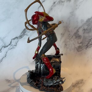 May include: A detailed figurine of a superhero in a dynamic pose, featuring a red and gold helmet and suit with black accents. The figure is mounted on a detailed base with metallic and dark brown tones, and includes multiple golden mechanical arms.
